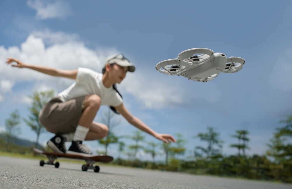 Osoba na deskorolce w tle, a na pierwszym planie unosi się biały dron z osłonami na śmigła. W tle widać drzewa i niebo.