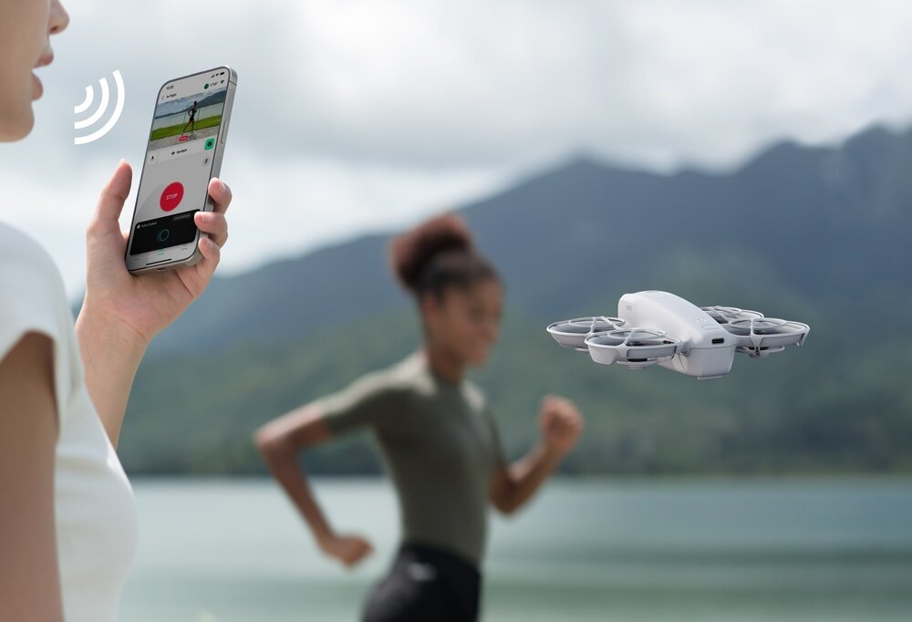 Osoba trzymająca smartfon z aplikacją do sterowania dronem, obok unosi się biały dron. W tle widoczna jest osoba biegnąca oraz krajobraz z górami i wodą.