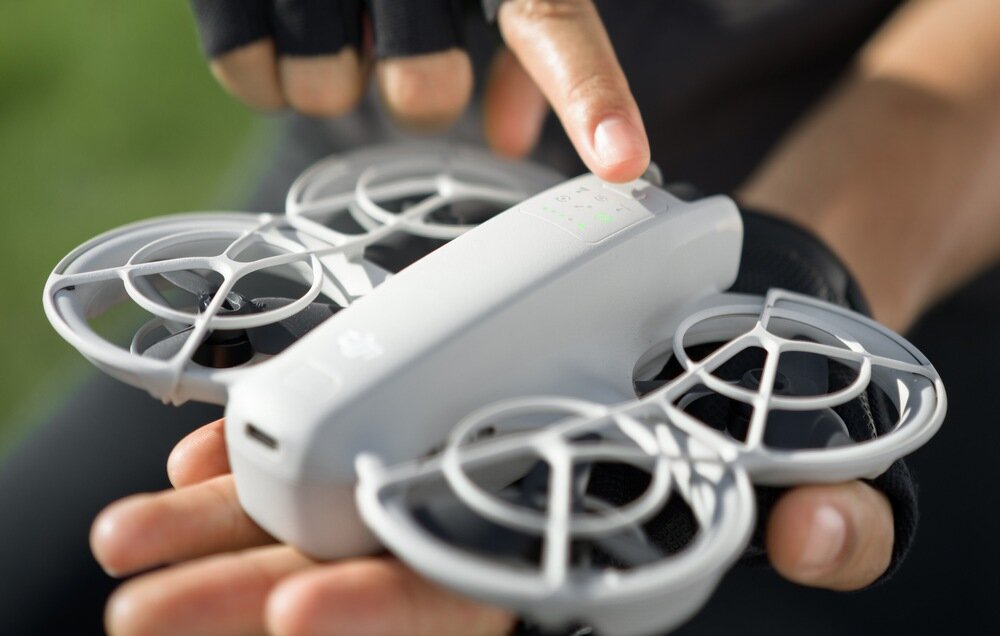 Osoba trzyma w dłoniach mały, biały dron z czterema osłoniętymi śmigłami. Na dronie widoczne jest logo DJI.
