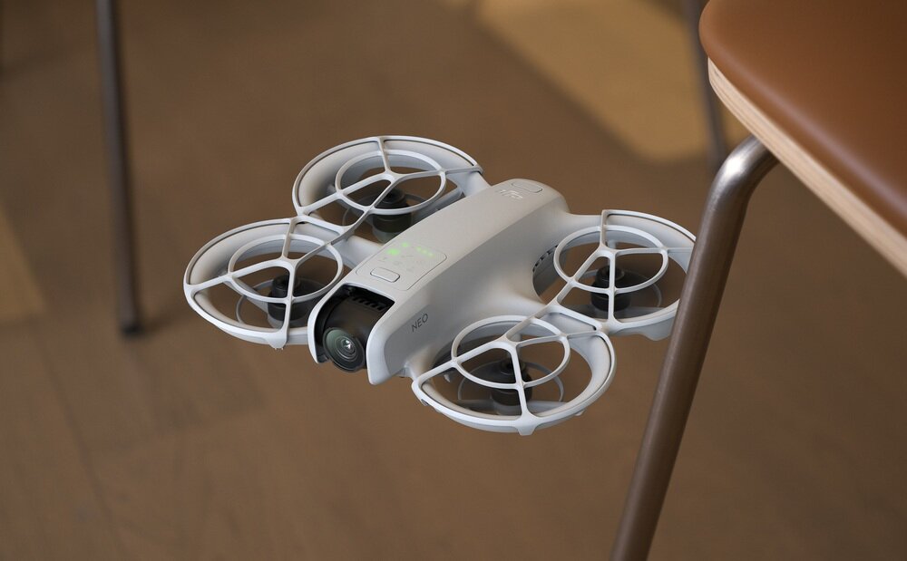 Na zdjęciu widać biały dron z czterema wirnikami, unoszący się w pomieszczeniu. Na obudowie drona widoczny jest napis \'NEO\'.