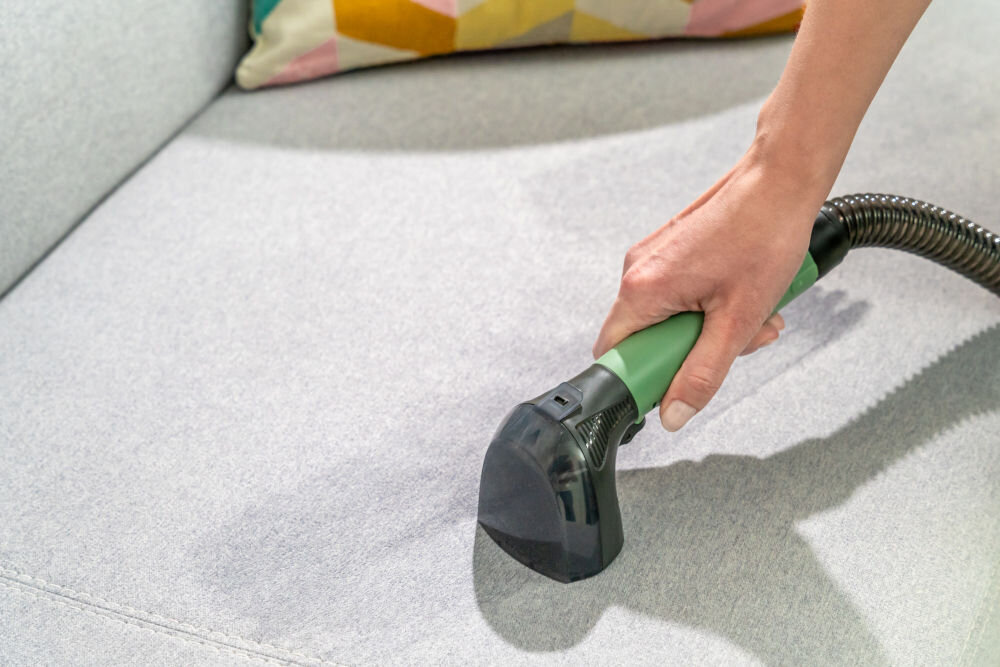 ODKURZACZ PIORĄCY SPOTCLEAN GOTZE&JENSEN CSC600 ze szczotką uniwersalną szeroką do butów do czyszczenia tapicerki meblowej samochodowej dywanów Zbliżenie na rękę kobiety używającej zielono-czarnej ssawki do czyszczenia jasnoszarej tapicerki sofy.