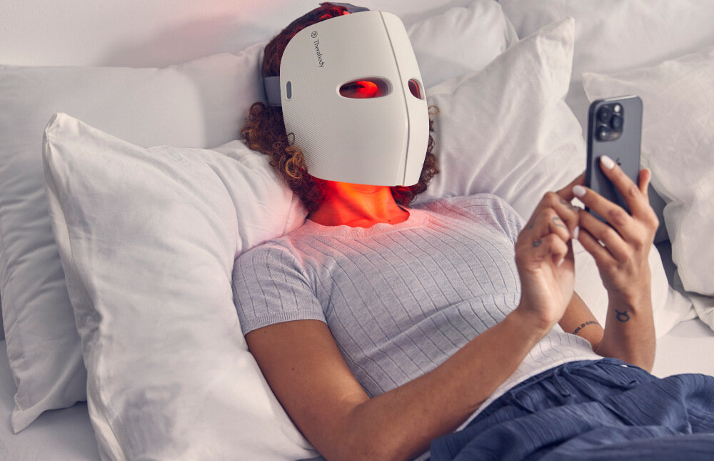Osoba leży na łóżku z białą pościelą, nosząc maskę LED na twarzy, która emituje czerwone światło. Trzyma w ręku smartfon, a na masce widoczne jest logo \'Therabody\'.