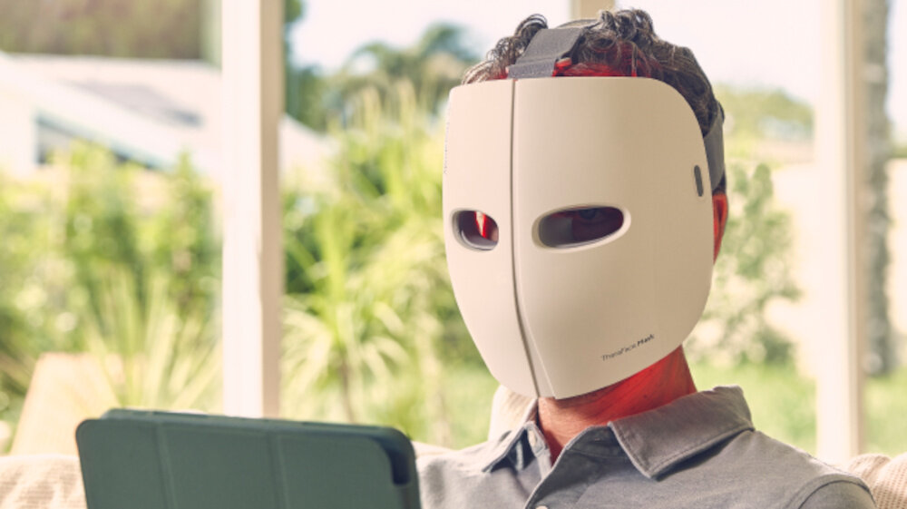 Osoba siedzi na kanapie, nosząc białą maskę LED na twarzy z dwoma otworami na oczy. W tle widać rozmyte rośliny i okno, a na masce znajduje się napis \'TheraFace Mask\'.