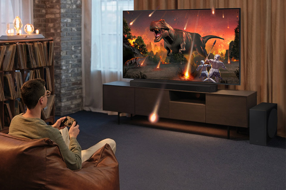 Osoba siedzi na kanapie i gra na konsoli, patrząc na duży telewizor, na którym wyświetlana jest scena z dinozaurem i meteorytami. Przed telewizorem znajduje się czarny soundbar i subwoofer.