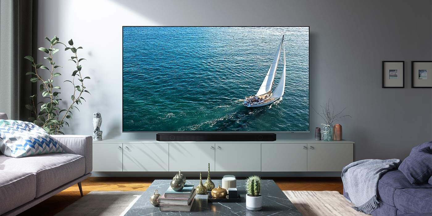 W salonie na szafce stoi duży telewizor wyświetlający obraz żaglówki na morzu, a pod nim znajduje się czarny soundbar. W pomieszczeniu widoczne są także kanapa, stolik z dekoracjami i kilka obrazków na ścianie.