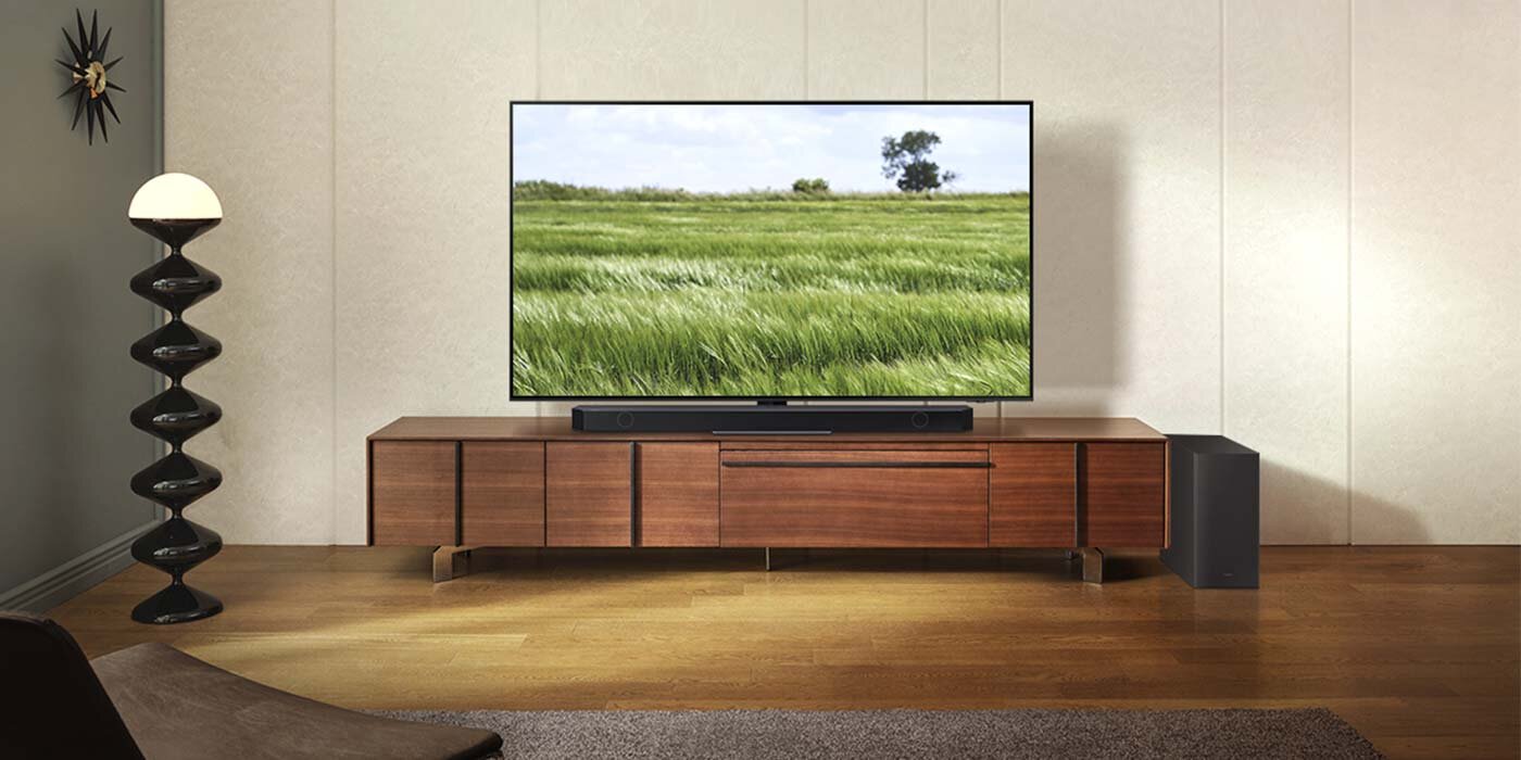 W salonie na drewnianej szafce stoi duży telewizor z wyświetlanym obrazem zielonego pola i drzewa. Przed telewizorem znajduje się czarny soundbar, a obok szafki stoi subwoofer.