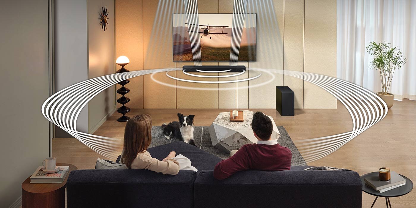 W salonie na kanapie siedzą dwie osoby, przed nimi znajduje się telewizor z wyświetlanym obrazem samolotu oraz soundbar. Wokół soundbara widoczne są graficzne fale dźwiękowe, a obok stoi subwoofer.