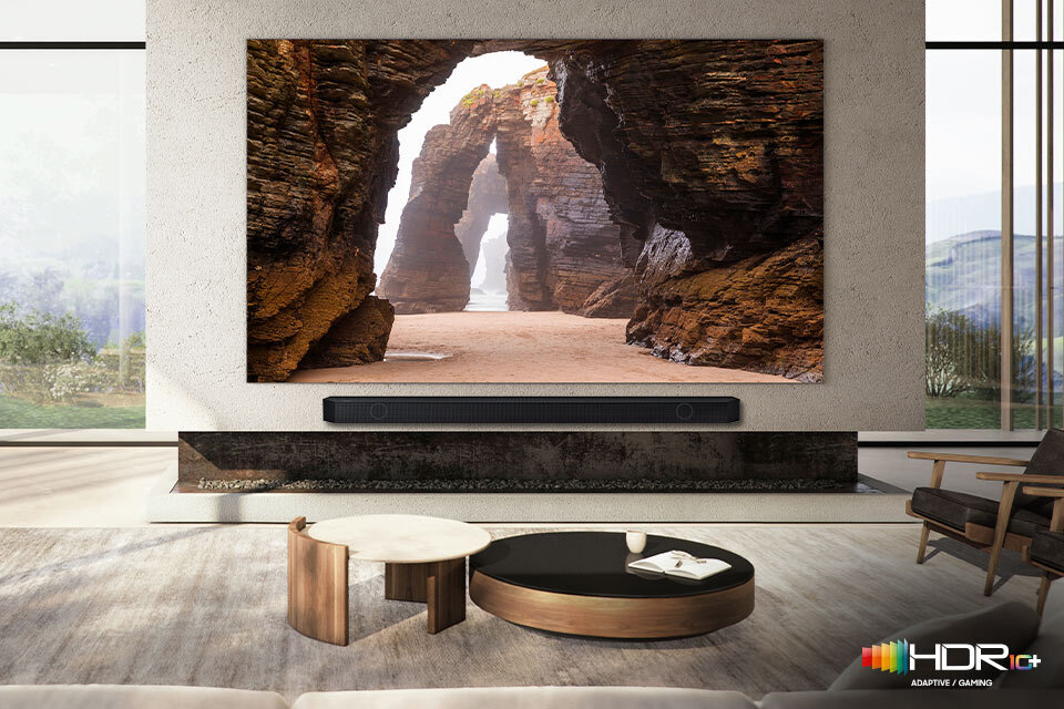 W salonie znajduje się duży telewizor wyświetlający obraz skalistego łuku nadmorskiego, a pod nim umieszczony jest czarny soundbar. W prawym dolnym rogu widoczne jest logo HDR10+ z napisem \'ADAPTIVE & GAMING\'.