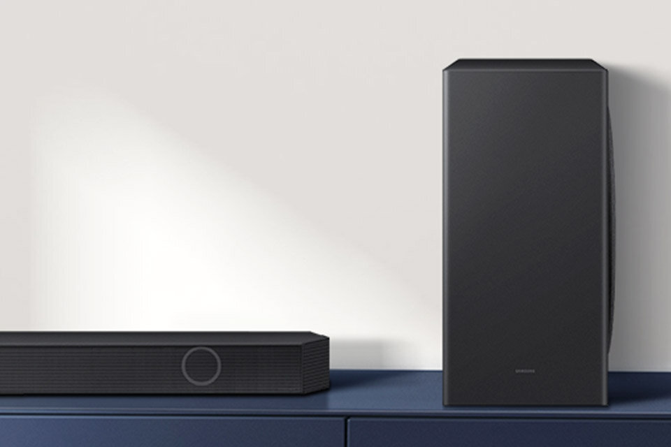 Na obrazie widoczny jest czarny soundbar oraz subwoofer ustawione na niebieskiej powierzchni. Na subwooferze znajduje się logo \'SAMSUNG\'.