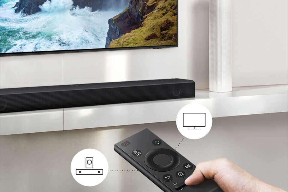 Na obrazie widać czarny soundbar umieszczony pod telewizorem, a w dłoni trzymany jest pilot z przyciskami. Obok pilota znajdują się dwie ikony: jedna przedstawiająca soundbar, a druga ekran.