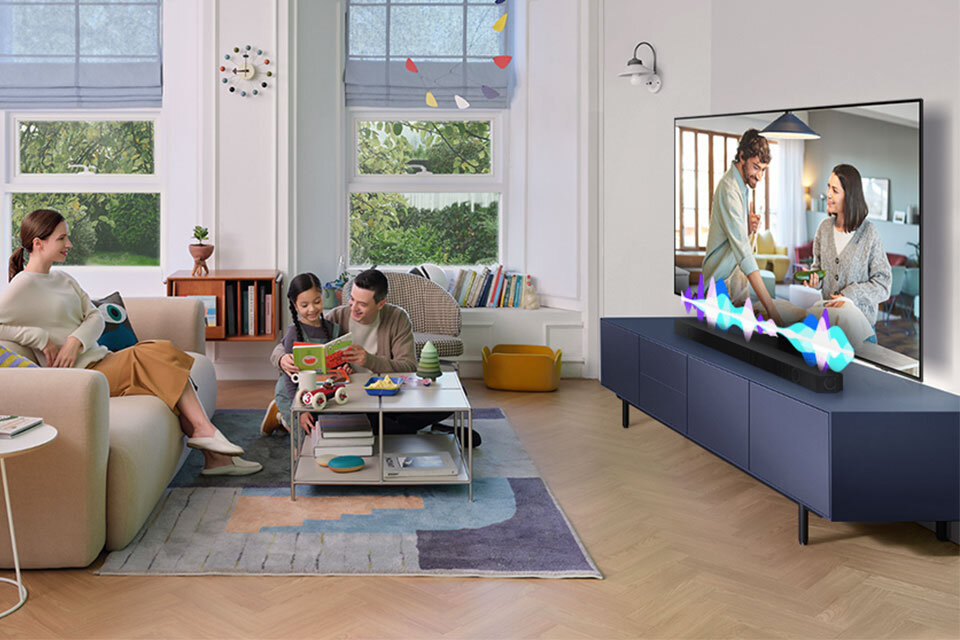 W salonie znajduje się telewizor na szafce, przed którym stoi soundbar. Na kanapie siedzą trzy osoby, a na ekranie telewizora widać dwie osoby rozmawiające.