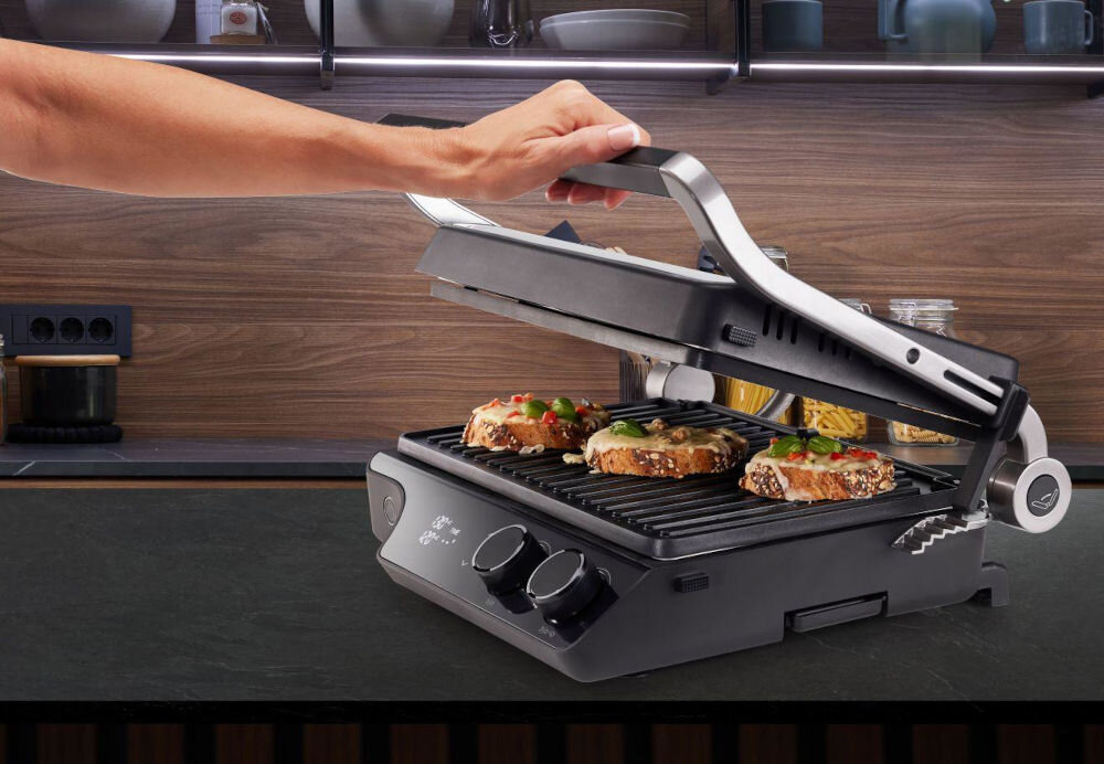 Osoba otwiera elektryczny grill, na którym znajdują się trzy kawałki pieczywa z dodatkami. W tle widać kuchenne półki z naczyniami i przyprawami.