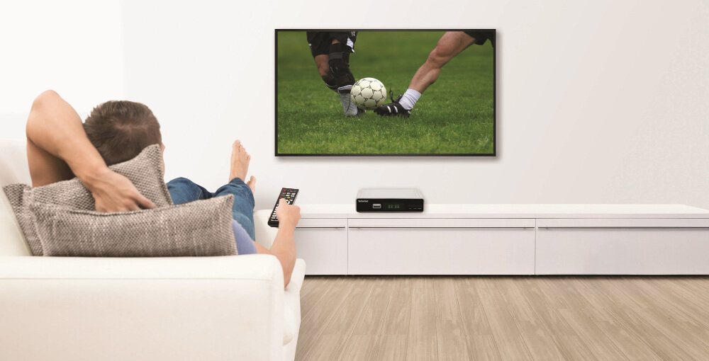 Osoba siedzi na kanapie i ogląda mecz piłki nożnej na telewizorze. Na szafce pod telewizorem znajduje się dekoder TECHNISAT.