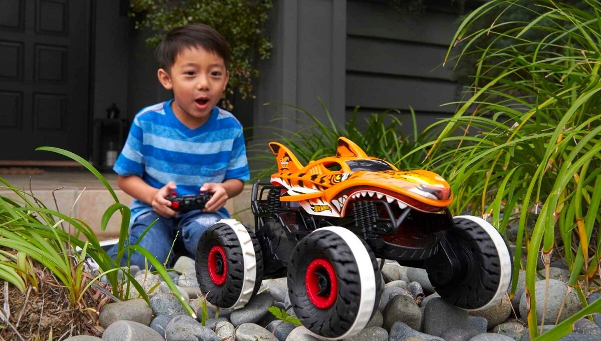 Dziecko bawi się zdalnie sterowanym samochodem terenowym o wyglądzie przypominającym rekina z pomarańczowym nadwoziem i dużymi kołami. W tle widoczne są rośliny i fragment budynku.