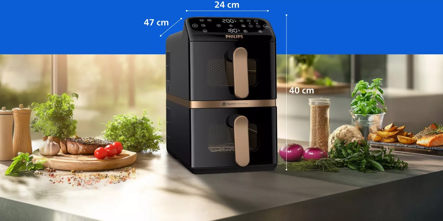 Na blacie kuchennym stoi czarna frytkownica beztłuszczowa z dwoma koszami, oznaczona logo PHILIPS. Obok urządzenia znajdują się świeże zioła, warzywa i przyprawy, a na ekranie frytkownicy widoczne są cyfry 200°C i 180°C.