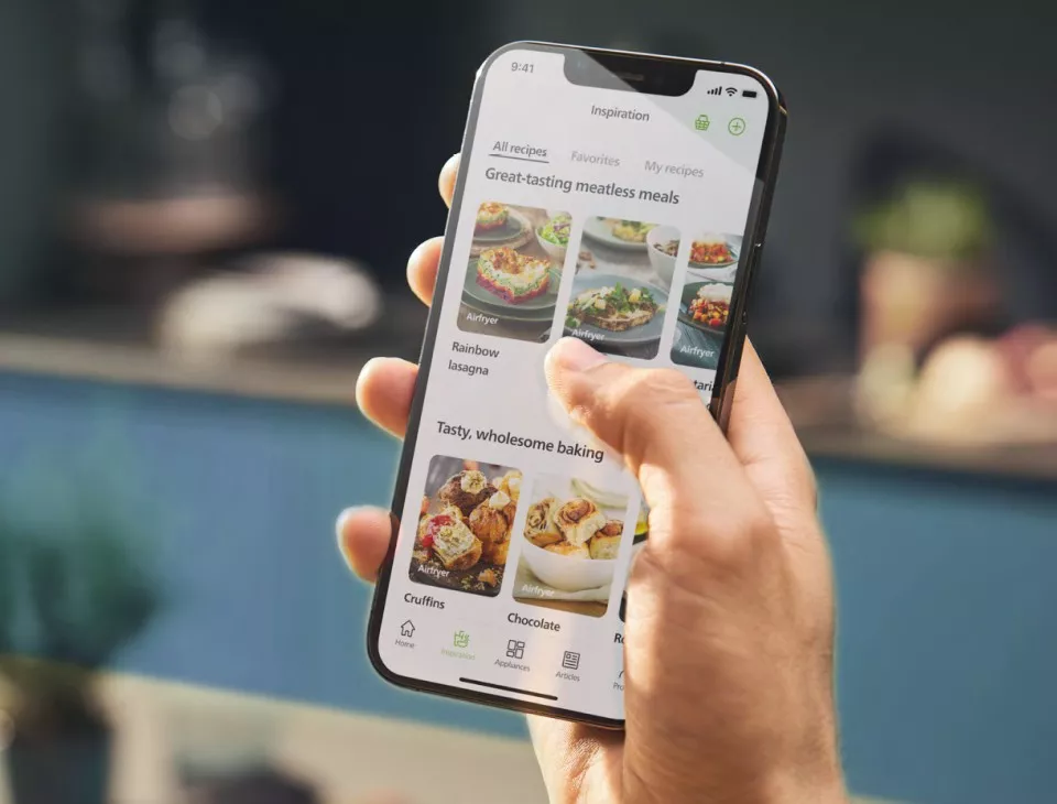 Osoba trzyma smartfon z aplikacją wyświetlającą przepisy kulinarne. Na ekranie widoczne są zdjęcia potraw oraz teksty takie jak \'Rainbow lasagna\' i \'Tasty, wholesome baking\'.