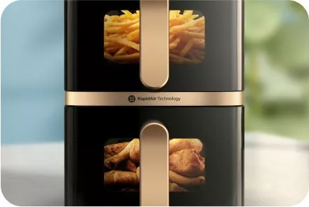 Obraz przedstawia dwie komory frytkownicy beztłuszczowej z przezroczystymi okienkami, w których widać frytki i kawałki mięsa. Na urządzeniu znajduje się napis \'Rapid Air Technology\'.