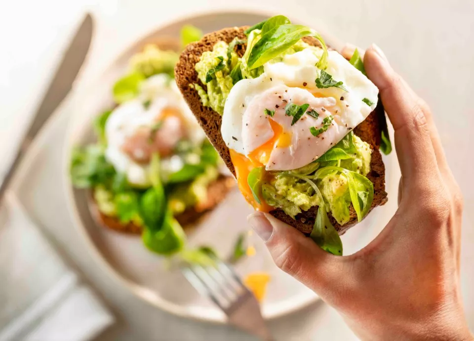 Osoba trzyma kromkę chleba z awokado, jajkiem w koszulce i zieleniną. W tle na talerzu znajduje się podobna kanapka oraz widelec.
