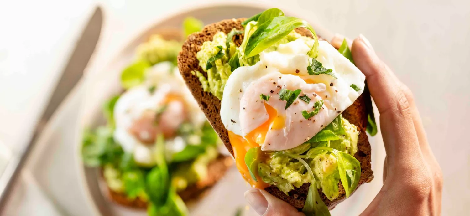 Osoba trzyma kromkę chleba z awokado, jajkiem w koszulce i świeżymi ziołami. W tle widoczny jest talerz z podobnie przygotowaną kanapką.