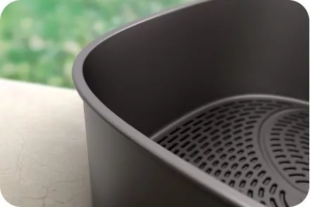 Zbliżenie na wnętrze metalowego kosza z perforowanym dnem, prawdopodobnie element frytkownicy. W tle widoczna jest rozmyta zielona powierzchnia.