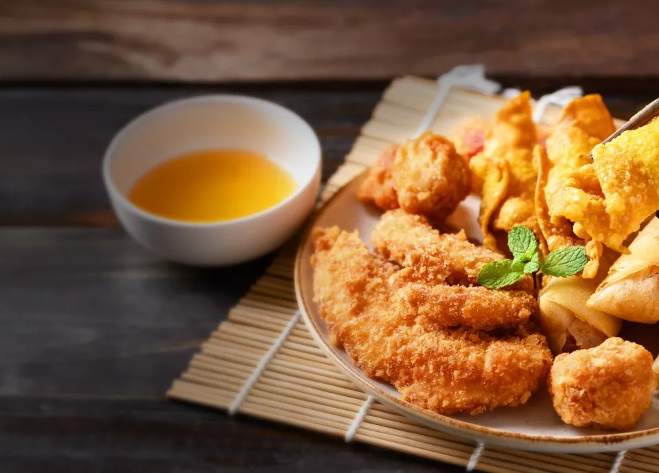 Na talerzu znajduje się różnorodność smażonych potraw, w tym panierowane kawałki mięsa i przekąski, ozdobione listkami mięty. Obok talerza stoi miseczka z jasnym sosem.