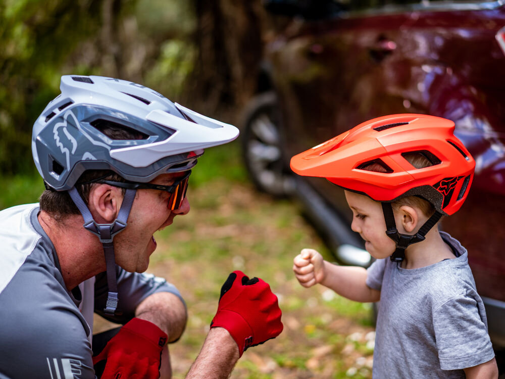 Na zdjęciu widać dwie osoby noszące kaski rowerowe, jedna w szarym, druga w czerwonym. W tle znajduje się samochód i zieleń.