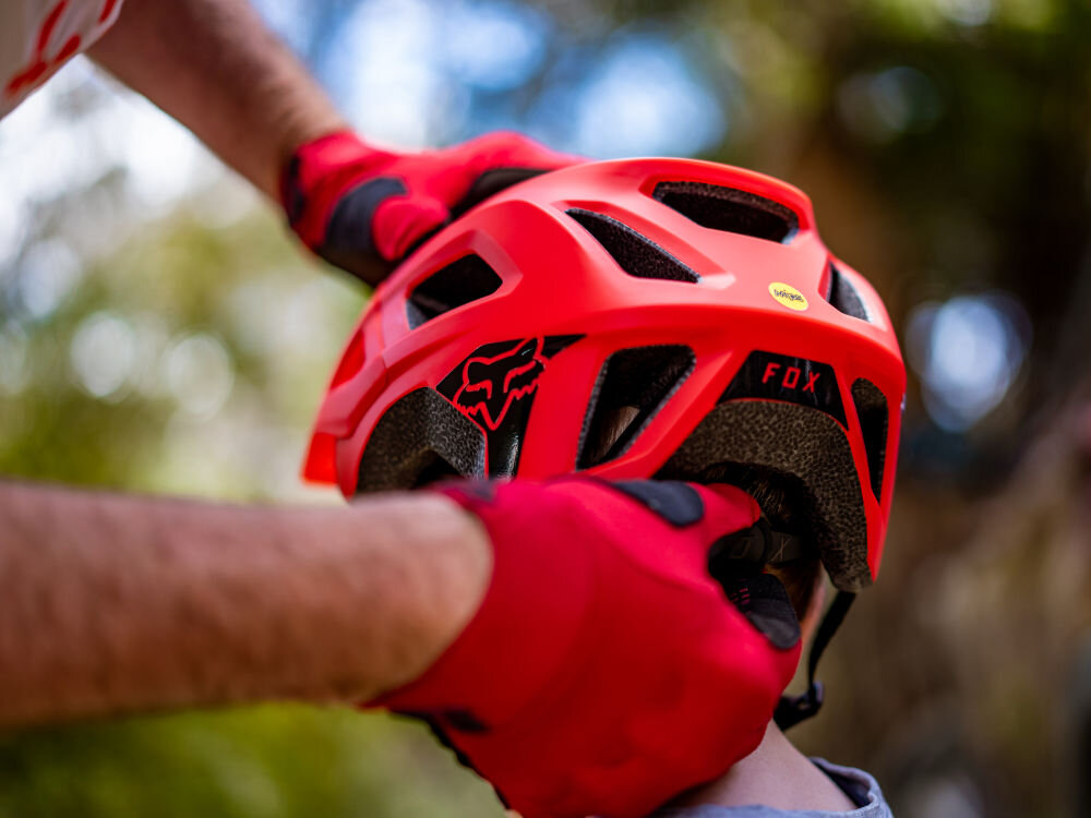 Czerwony kask rowerowy z widocznym logo \'FOX\' i żółtą naklejką \'MIPS\'. Osoba w czerwonych rękawiczkach trzyma kask na głowie innej osoby.