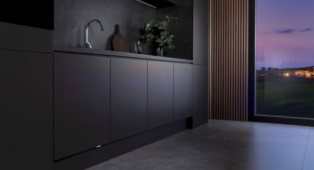Nowoczesna kuchnia z ciemnymi szafkami i zlewem, obok którego stoją dekoracje. Przez duże okno widać wieczorny krajobraz.