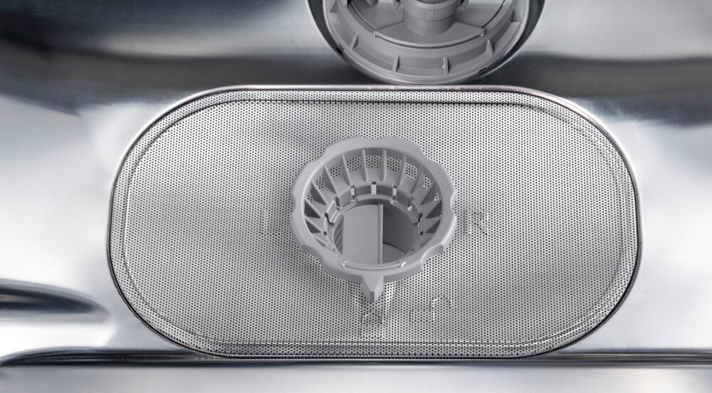Wnętrze zmywarki z widocznym metalowym filtrem i plastikowym elementem w kształcie koszyczka. Na powierzchni filtra widoczny jest tekst: \'FILTER\'.
