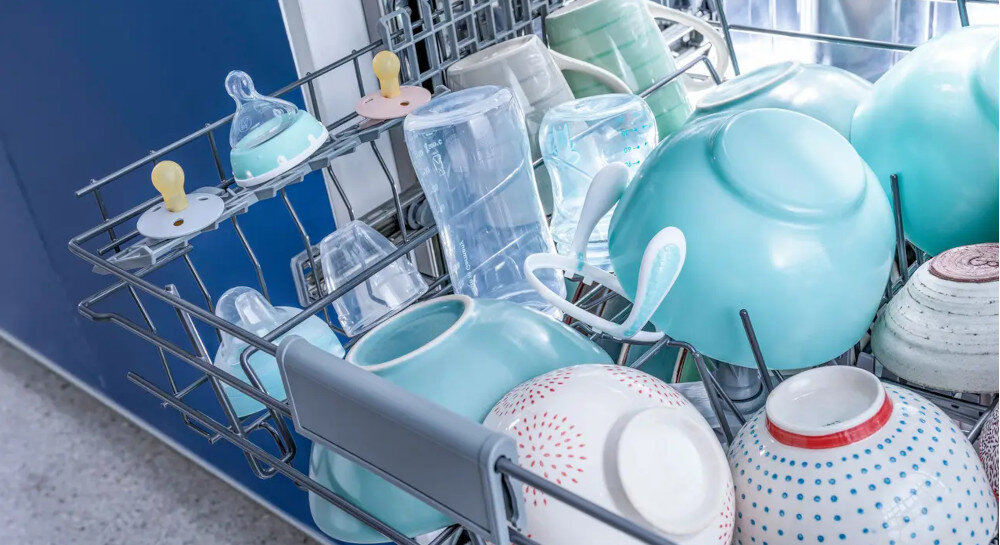 Wnętrze zmywarki z widocznymi naczyniami, w tym miski, kubki i butelki dla dzieci. Naczynia są ułożone w koszu zmywarki, a niektóre z nich są w pastelowych kolorach.