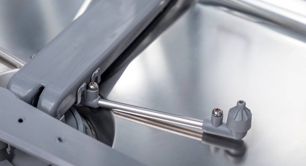 Zdjęcie przedstawia wnętrze zmywarki z widocznymi elementami mechanicznymi w kolorze szarym i metalicznym. Widać ramię spryskujące oraz metalowe rurki zamocowane śrubami.