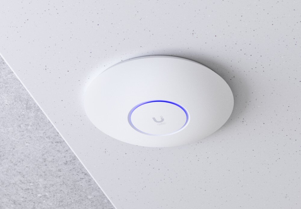 Punkt dostępu UBIQUITI UAP-AC-LR Białe urządzenie w kształcie dysku z niebieskim pierścieniem LED zamontowane płasko na jasnej powierzchni sufitu