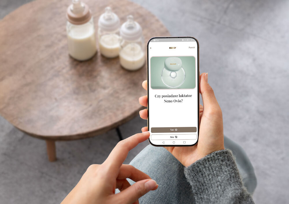 Osoba trzyma smartfon z aplikacją wyświetlającą pytanie \'Czy posiadasz laktator Neno Ovio?\' oraz dwie opcje odpowiedzi: \'Tak\' i \'Nie\'. W tle na stoliku znajdują się trzy butelki z mlekiem.