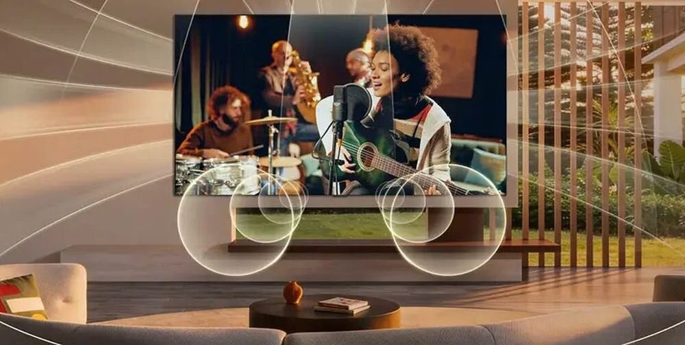 W salonie na ścianie wisi duży telewizor, na którym wyświetlany jest obraz zespołu muzycznego z wokalistką grającą na gitarze. Wokół telewizora widoczne są graficzne efekty dźwiękowe, sugerujące przestrzenny dźwięk.