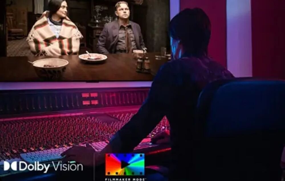 Osoba siedzi przed dużym ekranem, na którym wyświetlany jest film z dwiema postaciami przy stole. Na dole ekranu widoczne są loga \'Dolby Vision\' i \'Filmmaker Mode\'.