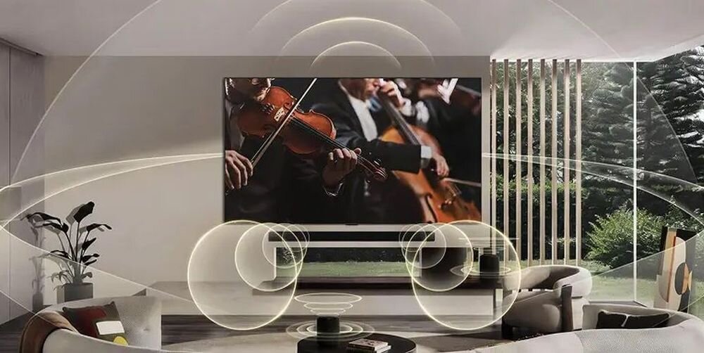W przestronnym salonie znajduje się duży telewizor wyświetlający obraz orkiestry grającej na instrumentach smyczkowych. Wokół telewizora i w pomieszczeniu widoczne są graficzne fale dźwiękowe, sugerujące efekt przestrzennego dźwięku.