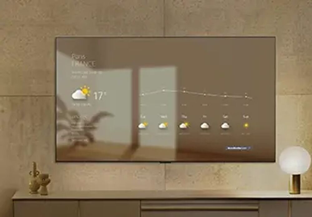 Na ścianie wisi duży telewizor, na którym wyświetlana jest prognoza pogody dla Paryża z temperaturą 17°C i ikoną słońca z chmurą. Obok telewizora znajduje się szafka z dekoracyjnymi przedmiotami i lampą.