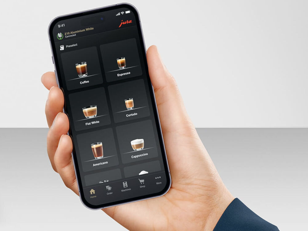 Osoba trzyma smartfon z aplikacją do wyboru napojów kawowych, takich jak Coffee, Espresso, Flat White, Cortado, Americano i Cappuccino. W górnym rogu ekranu widoczne jest logo \'jura\'.