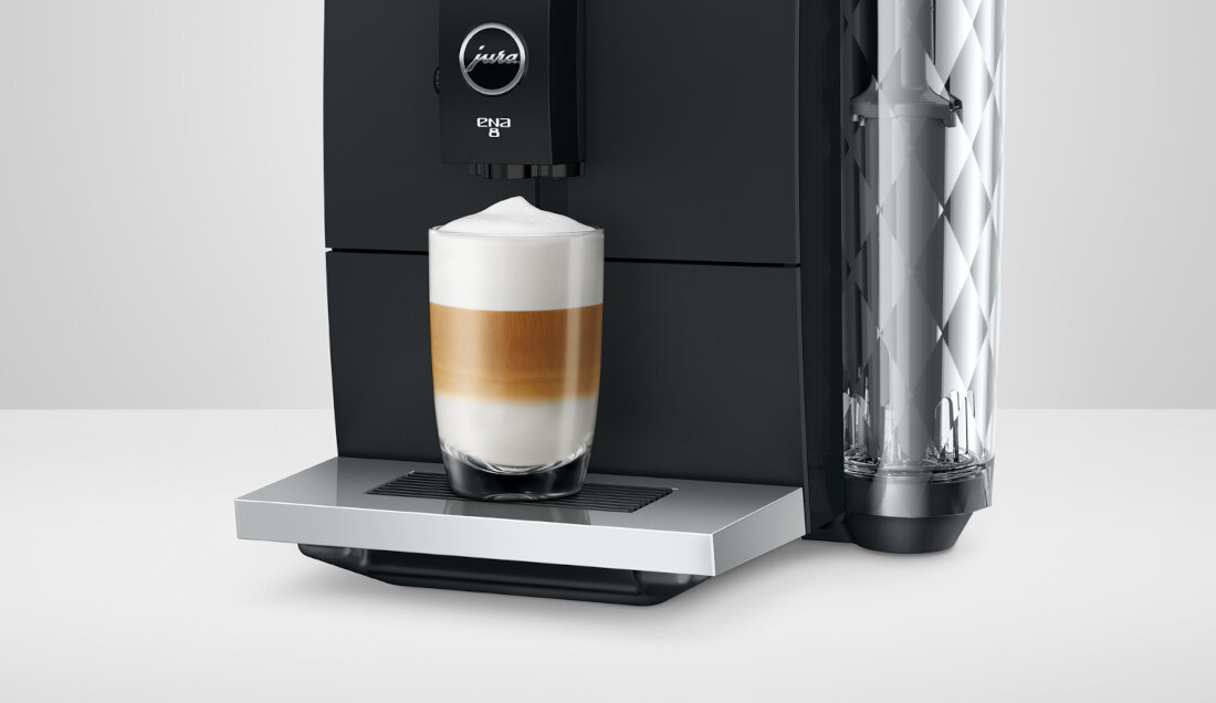 Czarny ekspres do kawy z logo \'jura\' i napisem \'ena 8\' przygotowuje kawę z mleczną pianką w szklance. Obok znajduje się przezroczysty pojemnik na wodę z diamentowym wzorem.