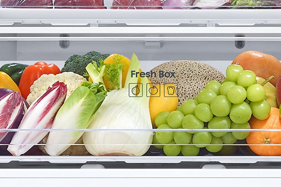 Wnętrze lodówki z szufladą oznaczoną jako \'Fresh Box\', wypełnioną różnorodnymi warzywami i owocami, takimi jak winogrona, pomarańcze, melon, brokuły i papryki. Na szufladzie widoczne są ikony przedstawiające owoce i warzywa.