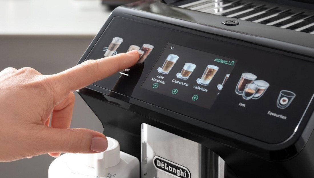 Osoba dotyka ekranu dotykowego ekspresu do kawy, na którym wyświetlane są różne opcje napojów, takie jak Latte Macchiato, Cappuccino i Caffè latte. Na urządzeniu widoczne jest logo \'DeLonghi\'.