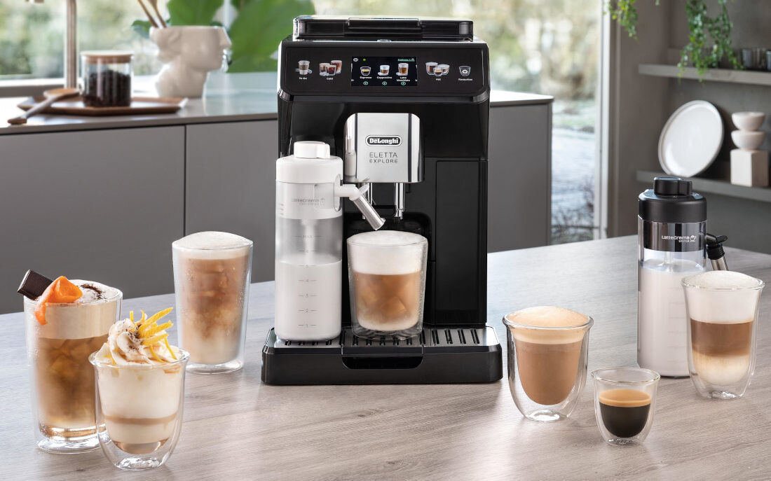 Na blacie kuchennym stoi ekspres do kawy z logo \'DeLonghi Eletta Explore\', otoczony różnymi filiżankami z kawą. Obok ekspresu znajduje się pojemnik na mleko oraz kilka szklanek z różnymi napojami kawowymi.