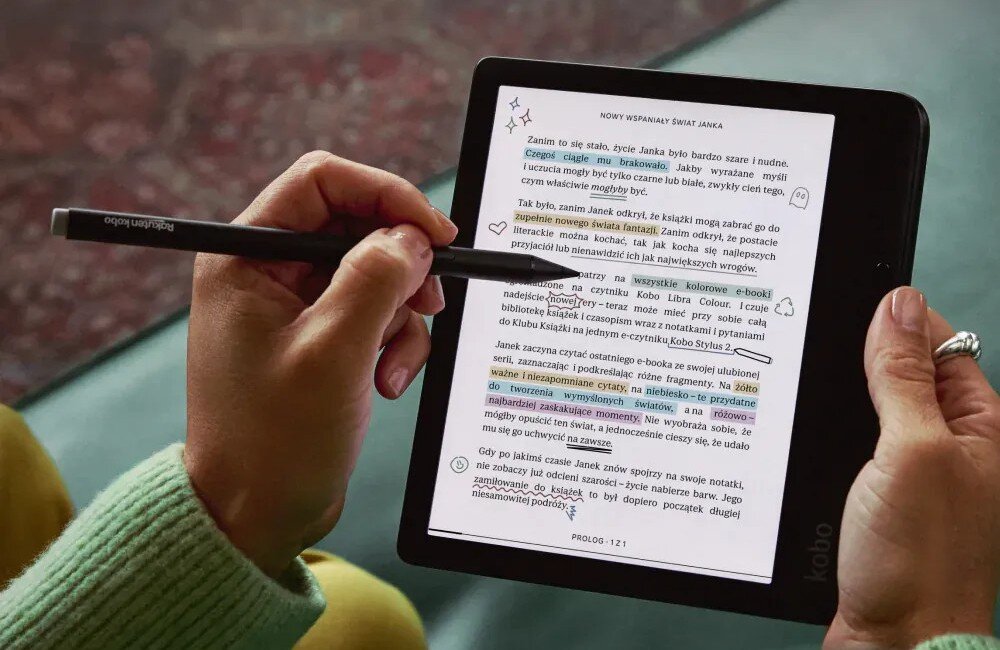 Osoba trzyma czytnik e-booków KOBO, na którym widoczny jest tekst z podkreśleniami i notatkami. Na ekranie znajduje się fragment tekstu zatytułowany \'NOWY WSPANIAŁY ŚWIAT JANKA\'.