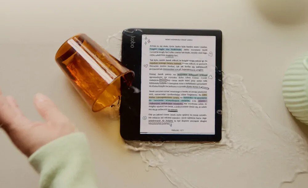 Na stole leży czytnik e-booków z widocznym tekstem na ekranie, obok przewrócony kubek z rozlaną cieczą. Na urządzeniu widoczne jest logo \'kobo\'.