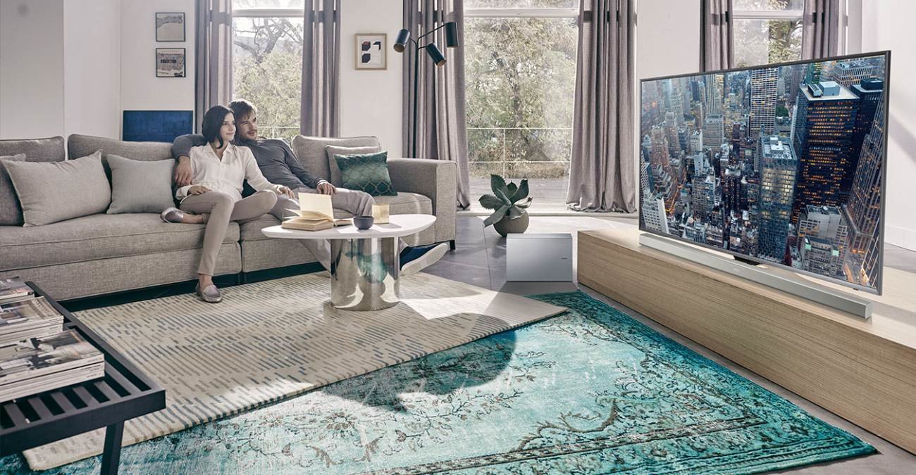 Zdjęcie przedstawiające kobietę i mężczyznę siedzących na kanapie w nowoczesnym mieszkaniu. Patrzą na telewizor Samsung, pod którym stoi płaski Soundbar Samsung w kolorze szarym.