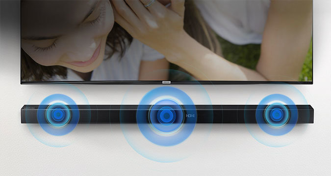 Ilustracja przedstawiająca płaski Samsung Soundbar w kolorze czarnym, zawieszony pod wiszącym telewizorem Samsung. Z głośników Soundbara wydobywa się dźwięk. Telewizor wyświetla uśmiechniętą kobietę.