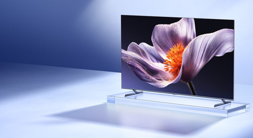 Telewizor z cienką ramką wyświetla obraz dużego, fioletowego kwiatu z pomarańczowym środkiem. Urządzenie stoi na przezroczystej podstawie, a tło jest w odcieniach niebieskiego.