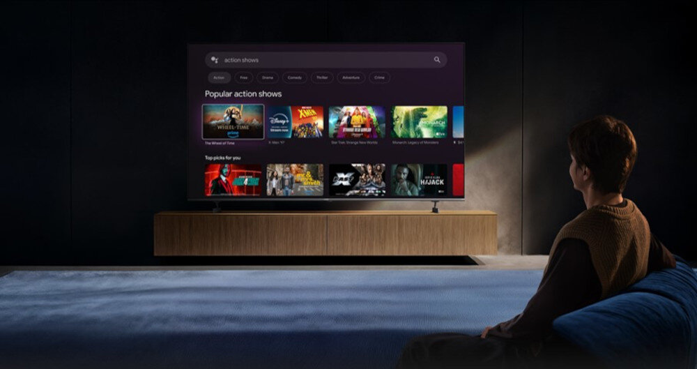 Osoba siedzi na kanapie przed dużym telewizorem, na którym wyświetlane jest menu z popularnymi programami akcji. Na ekranie widoczne są miniatury różnych filmów i seriali.