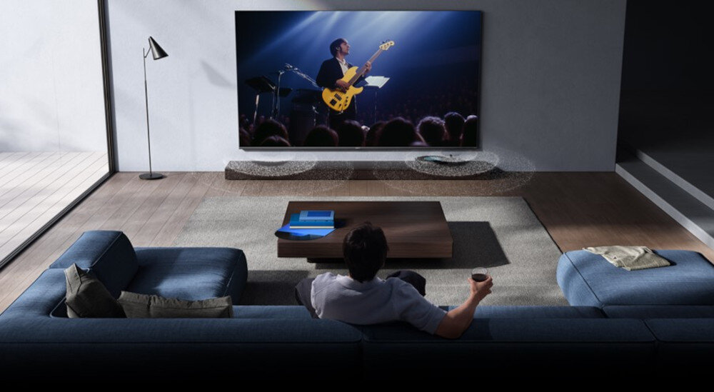 Osoba siedzi na kanapie w salonie, oglądając duży telewizor, na którym wyświetlany jest koncert muzyczny. W pomieszczeniu znajduje się również stolik kawowy i lampa stojąca.