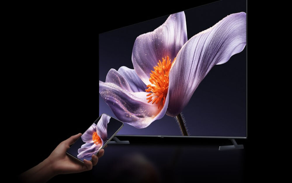 Obraz przedstawia duży telewizor z wyświetlonym na ekranie zbliżeniem fioletowego kwiatu z pomarańczowym środkiem. Przed telewizorem znajduje się ręka trzymająca smartfon, na którym widoczny jest ten sam obraz kwiatu.